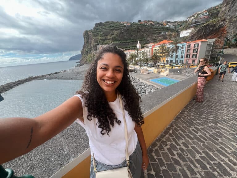 Is Madeira Worth Visiting? An Honest Review 1 Woman smiling and taking a selfie along the waterfront in Ponta do Sol, Madeira, with colorful buildings, palm trees, and steep cliffs along the coast behind her.